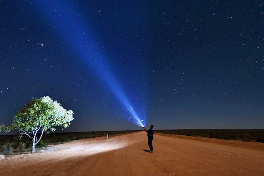 A person stands holding a torch looking up at the night sky which is filled with stars.