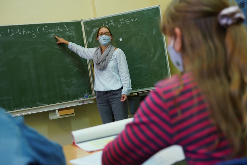 A teacher wears a mask as she points to a blackboard. Young girl in foreground also wearing mask.