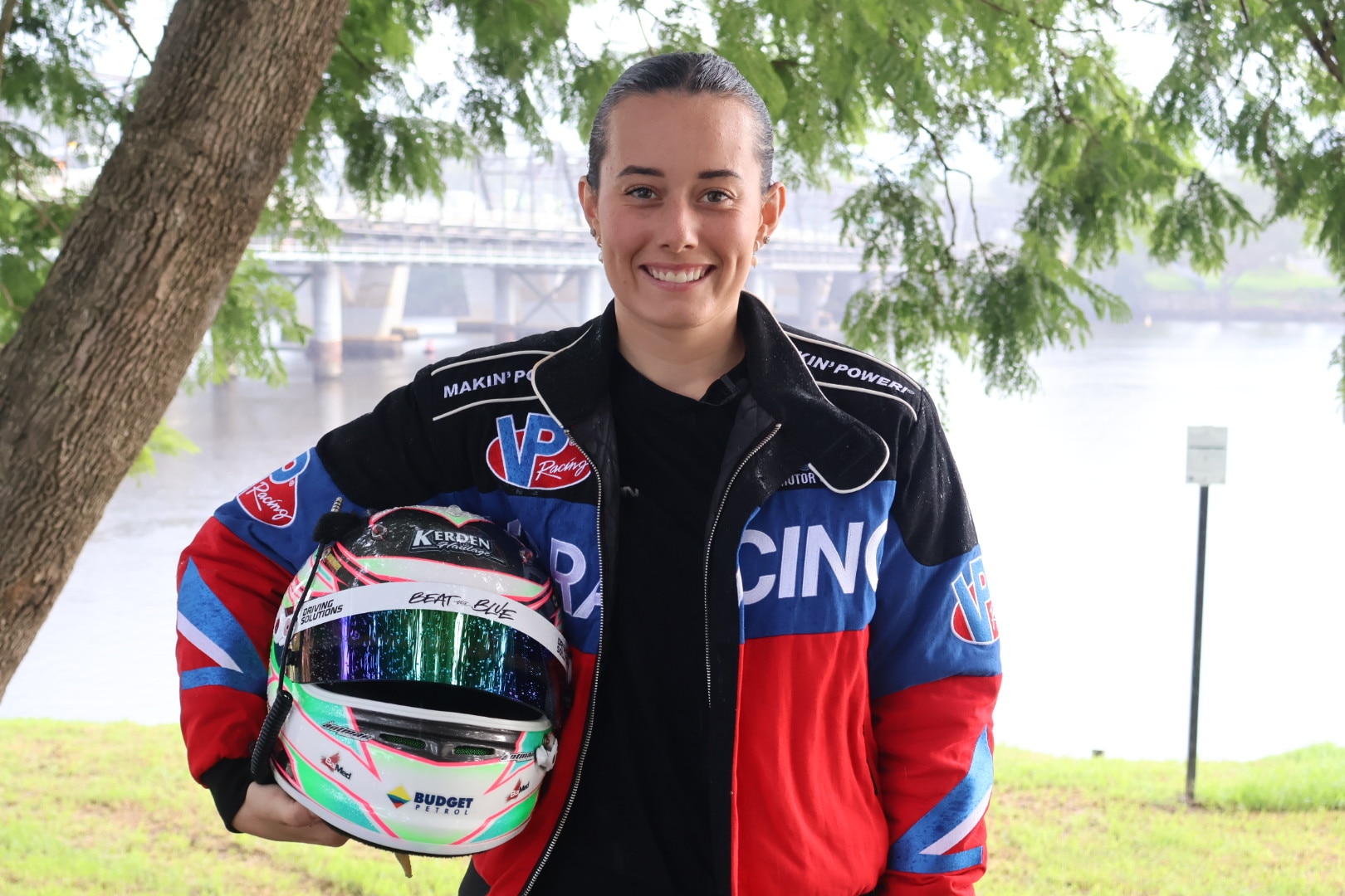 Imogen veste uma jaqueta vermelha e azul e segura o capacete, sorrindo com o rio atrás dela.