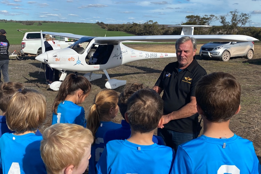 Back of schhool children in blue uniforms heads, man talking to them, small plane in background