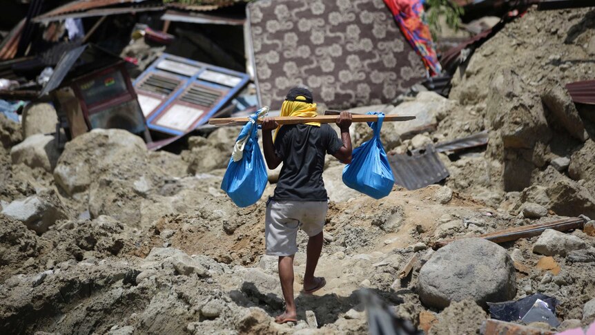 a man carries belongings from his toppled house. He balances two blue bags on a piece of wood on his shoulders.