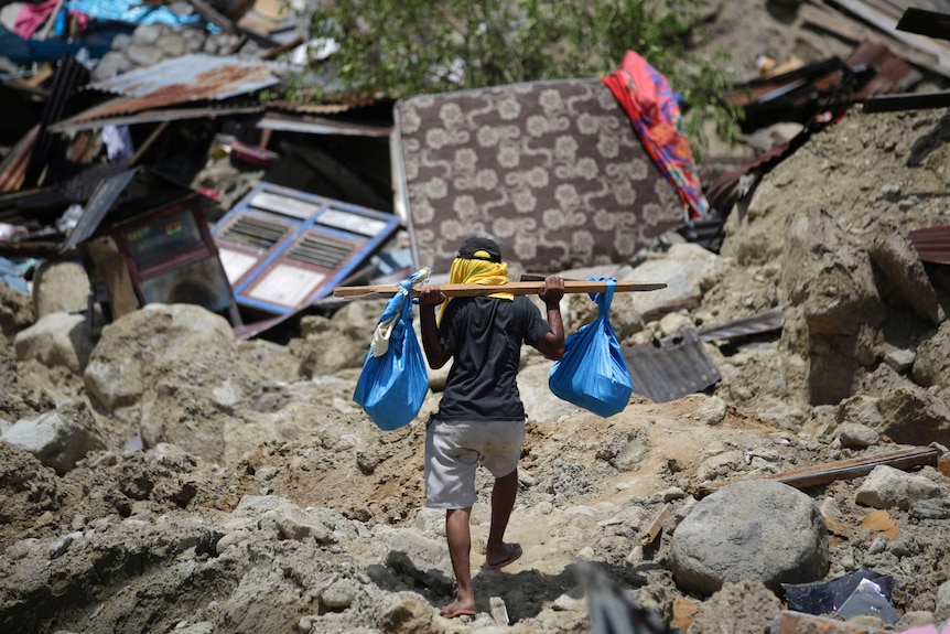 A man carries belongings from his toppled house. He balances two blue bags on a piece of wood on his shoulders.