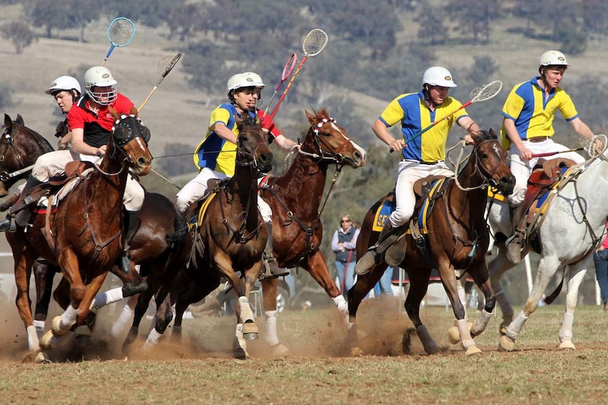 Six helmet-wearing players ride horses and hold rackets in the air as dust flies beneath the horses' hooves and spectators watch