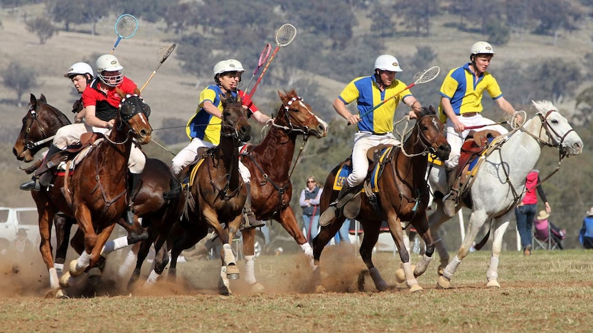 Six helmet-wearing players ride horses and hold rackets in the air as dust flies beneath the horses' hooves and spectators watch