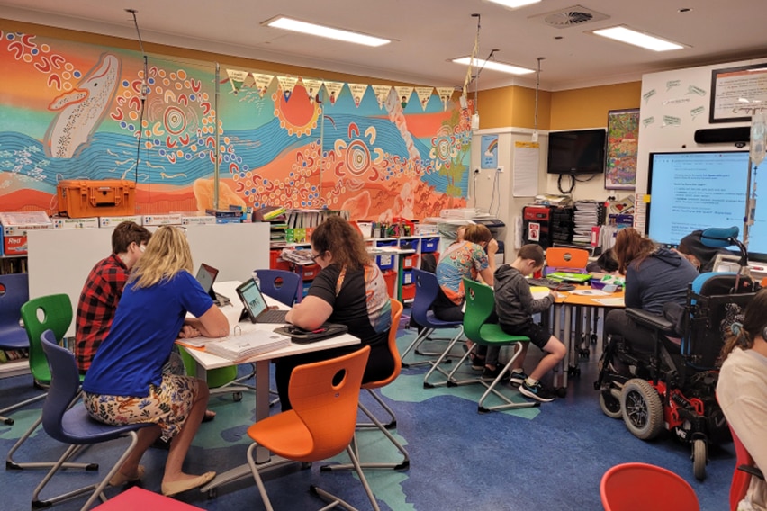 Students and teachers sitting at two separate desks in a small colourful classroom 