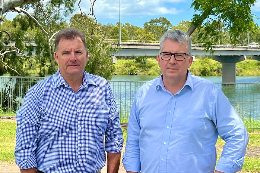 Photo of two middle-aged white men standing in front of a bridge, surrounded by green trees. 