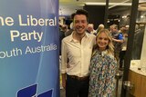 A young Liberal man stands with a woman in front of a Liberal Party sign.