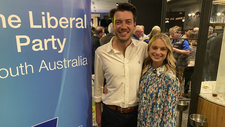 A young Liberal man stands with a woman in front of a Liberal Party sign.