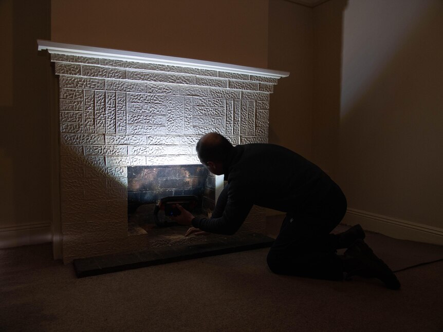 A man crouches down in front of a fireplace, with a torch pointed up the chimney
