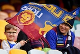 A young Brisbane Lions fan waves a flag as he smiles in the stands at the Gabba next to two adults.