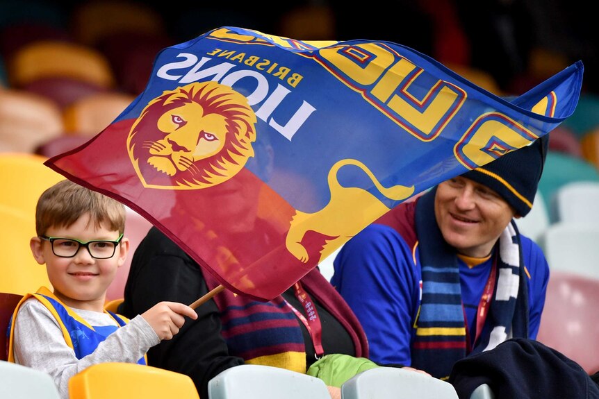 A young Brisbane Lions fan waves a flag as he smiles in the stands at the Gabba next to two adults.