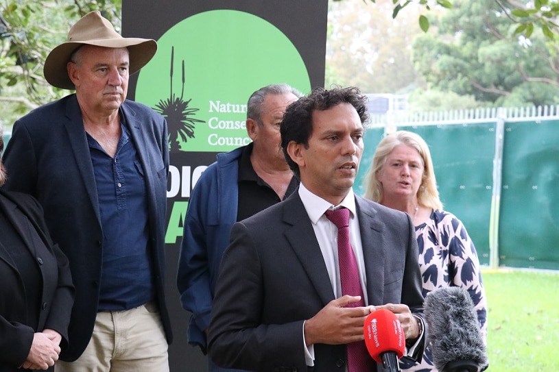 A group of people stand behind a man in a suit making a speech.