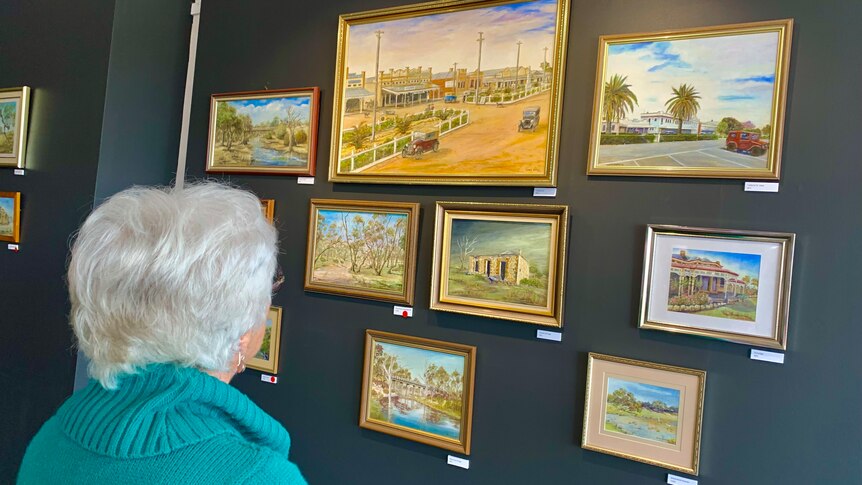 A woman with grey hair and her back to the camera admires landscape paintings on a grey charcoal wall