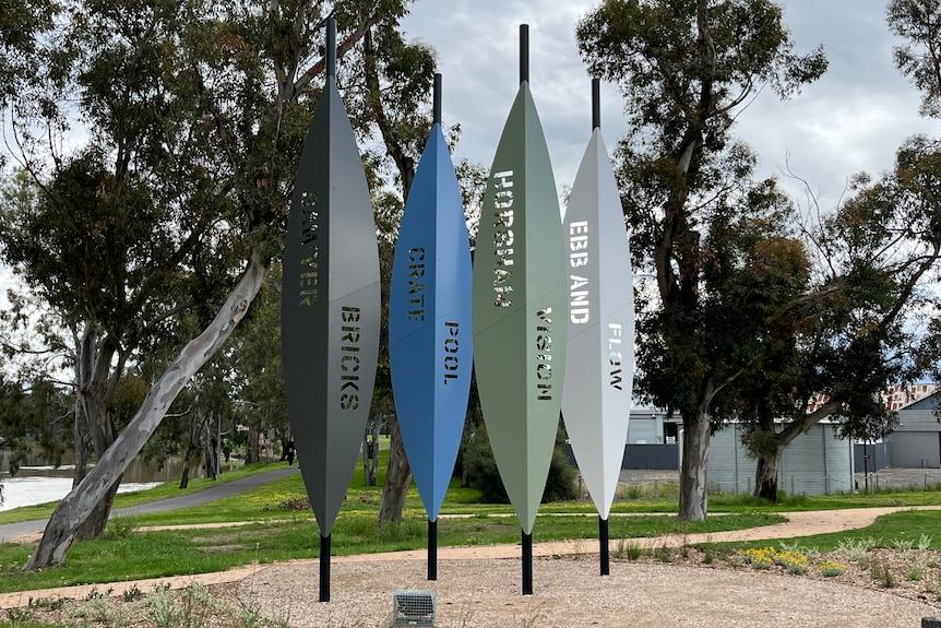 A sculpture of gum-leaf shaped, metal fins in black, blue, green and grey against the backdrop of the river and park.