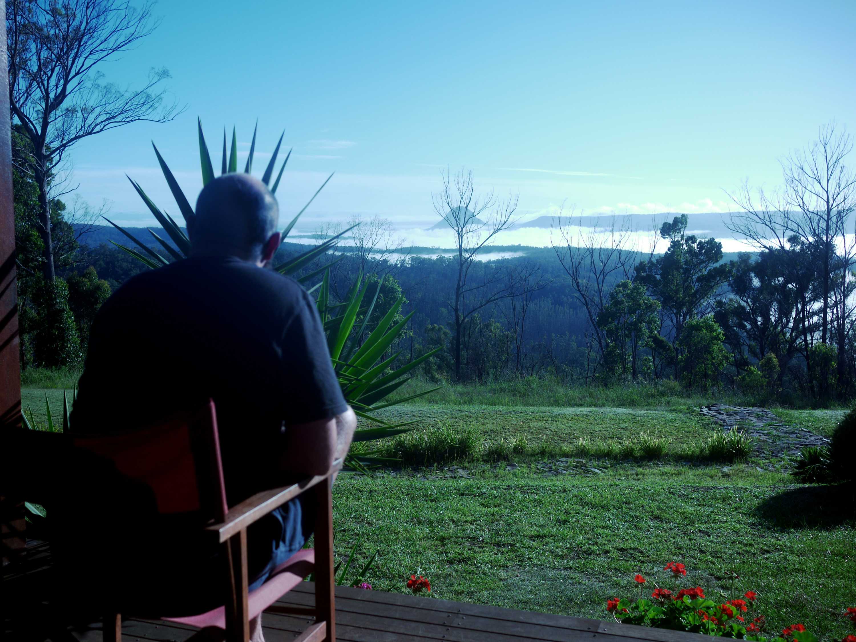 A man is seen from behind sitting on the deck of a house and looking a view of mountains in the distance.