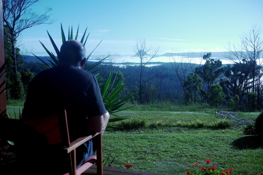 A man is seen from behind sitting on the deck of a house and looking a view of mountains in the distance.