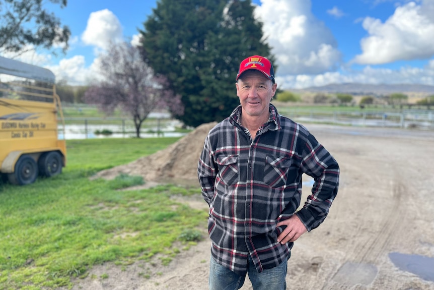 A man in a plaid shirt and a red cap standing in front of a driveway with a horse float to the side.