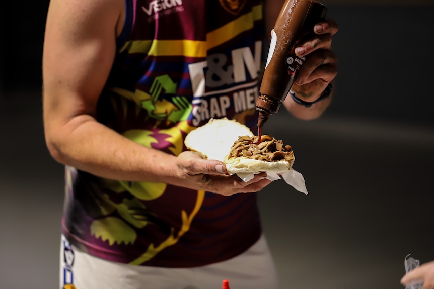 A bottle of barbecue sauce is squeezed onto a lamb roll by a young man wearing a singlet and shorts