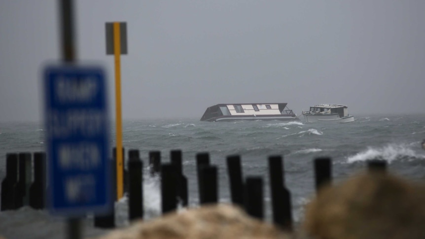 A houseboat lies partly submerged in choppy water under grey skies, with timber pylons in the foreground.