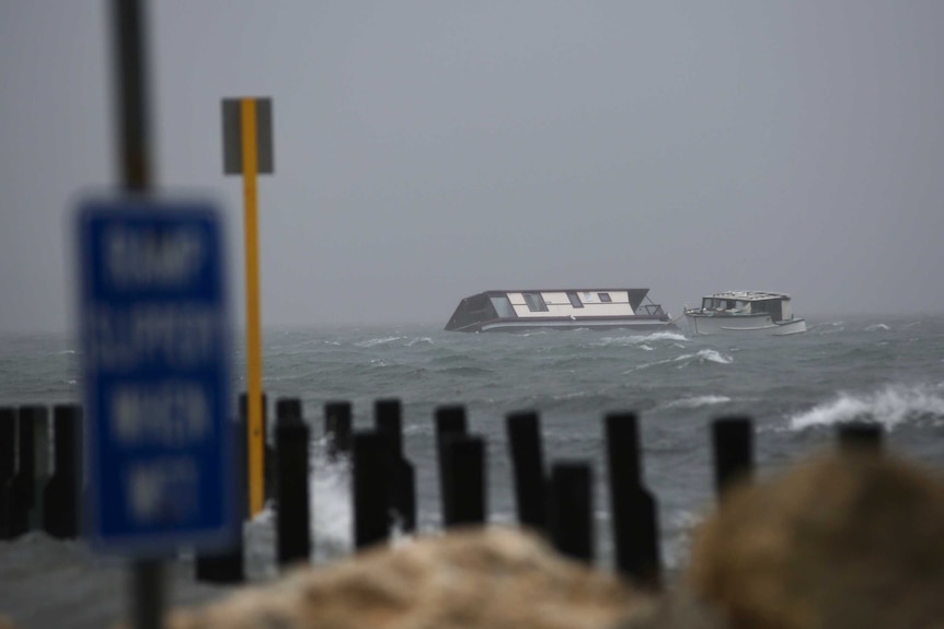 A houseboat lies partly submerged in choppy water under grey skies, with timber pylons in the foreground.