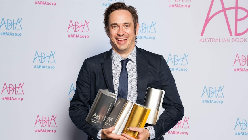 A man in a navy jacket and tie stands against a white media wall smilling and holding a handful of trophies.