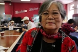 A woman wearing large round framed glasses smiles neutrally while sitting in a cafeteria