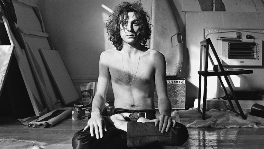 A black-and-white photo of Pink Floyd's Syd Barrett sitting cross-legged on the floor, with no shirt on