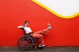 Tessa Deak sits in her wheelchair wearing a white top and pinkish-red pants, while holding one leg up to show her rainbow shoes.