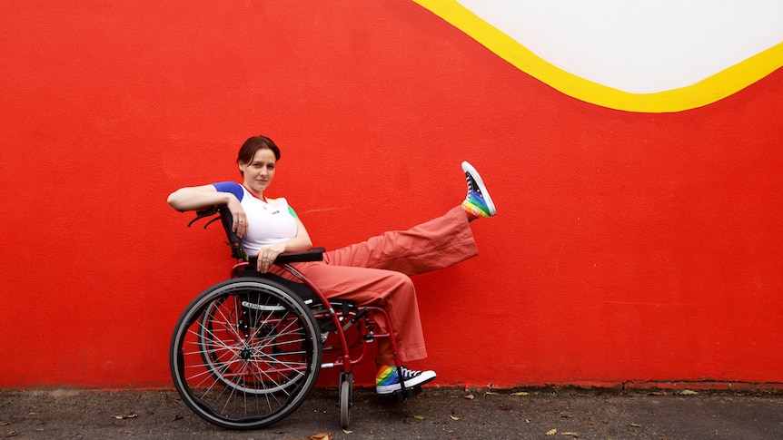 Tessa Deak sits in her wheelchair wearing a white top and pinkish-red pants, while holding one leg up to show her rainbow shoes.