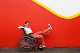 Tessa Deak sits in her wheelchair wearing a white top and pinkish-red pants, while holding one leg up to show her rainbow shoes.
