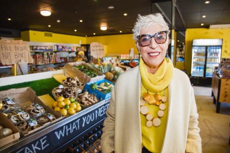 An older woman wearing yellow stands in a fresh food market.