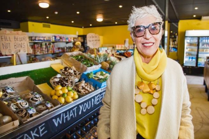 An older woman wearing yellow stands in a fresh food market.