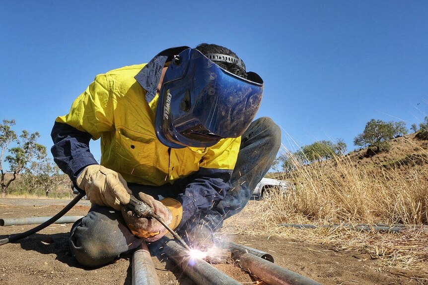 A man crouching and welding metal fence posts together.