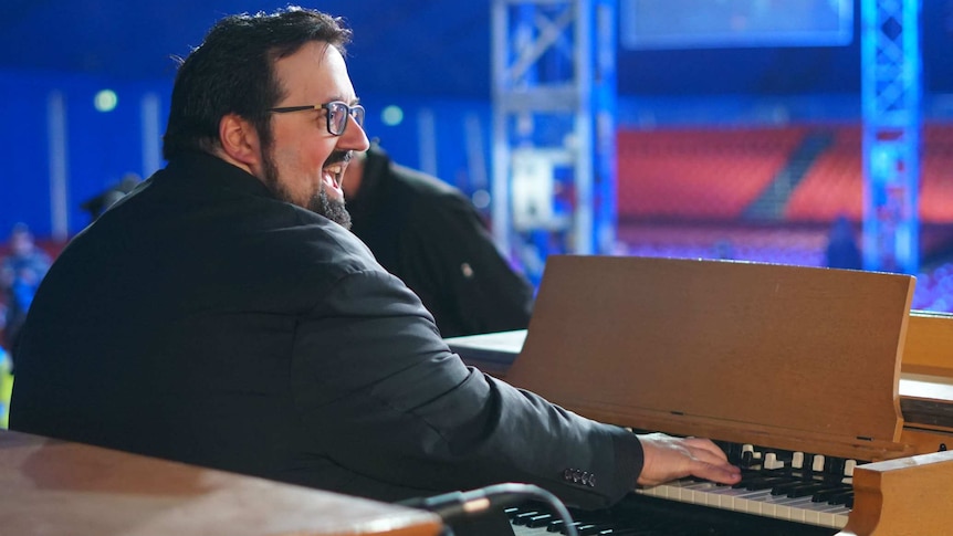 A man in a black jacket, wearing glasses, with dark brown hair, smiles as he plays an organ.