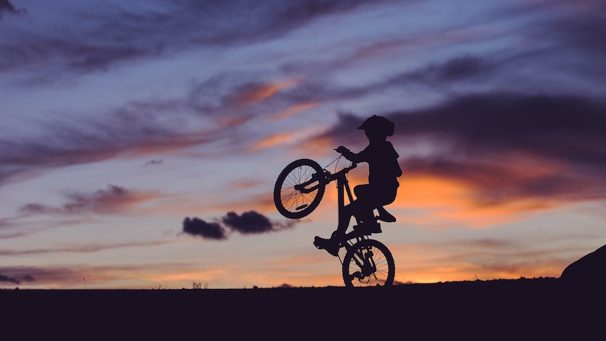 Silhouette image of a boy tipping his bike on the back wheel as the sun goes down