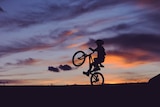Silhouette image of a boy tipping his bike on the back wheel as the sun goes down