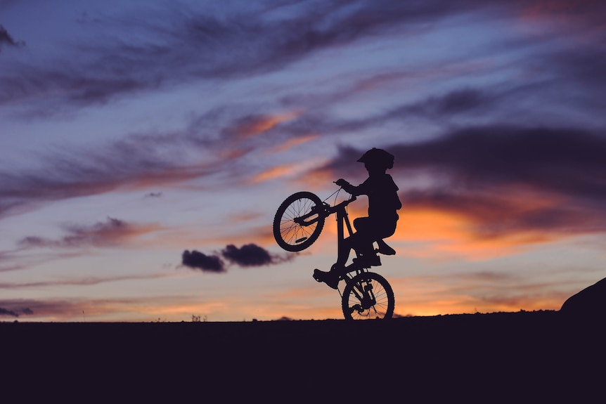 Silhouette image of a boy tipping his bike on the back wheel as the sun goes down