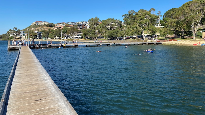 A scenic photo of a jetty near a stretch of coastline with people on it fishing and swimming.