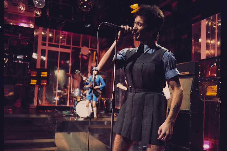 Bon Scott wears schoolgirl drag and wig on stage next to guitarist Angus Young in his trademark schoolboy outfit.