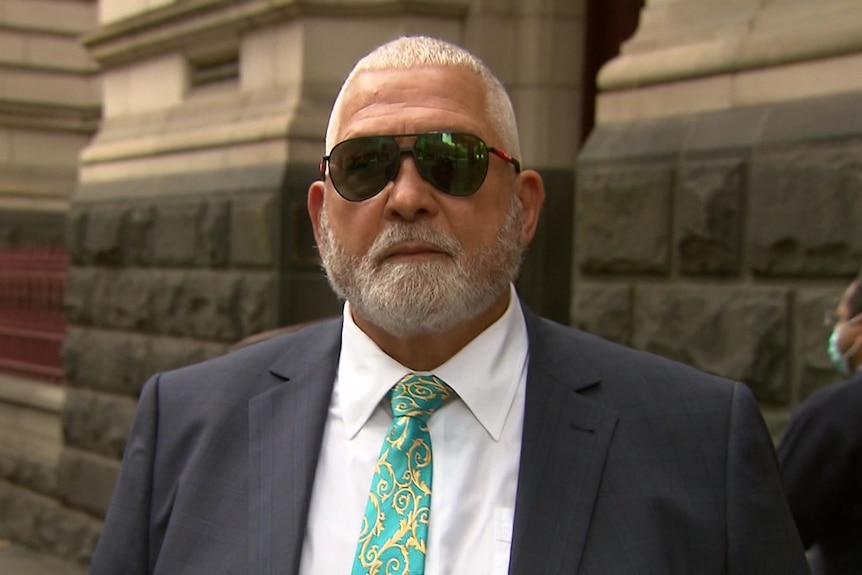Un homme portant des lunettes de soleil et un costume-cravate se tient devant un bâtiment officiel.