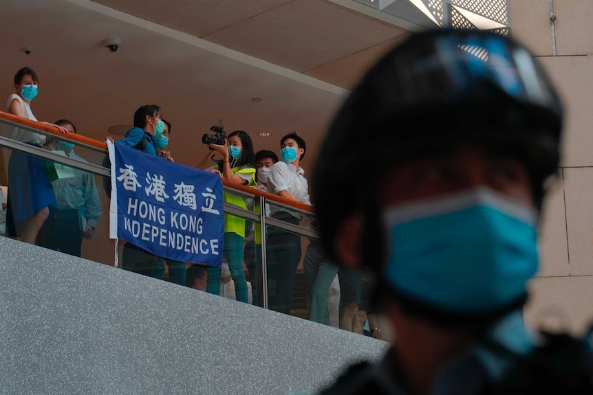 A police officer in the foreground with a balcony in the background are people in masks with a sign saying "Hong Kong Indepence"