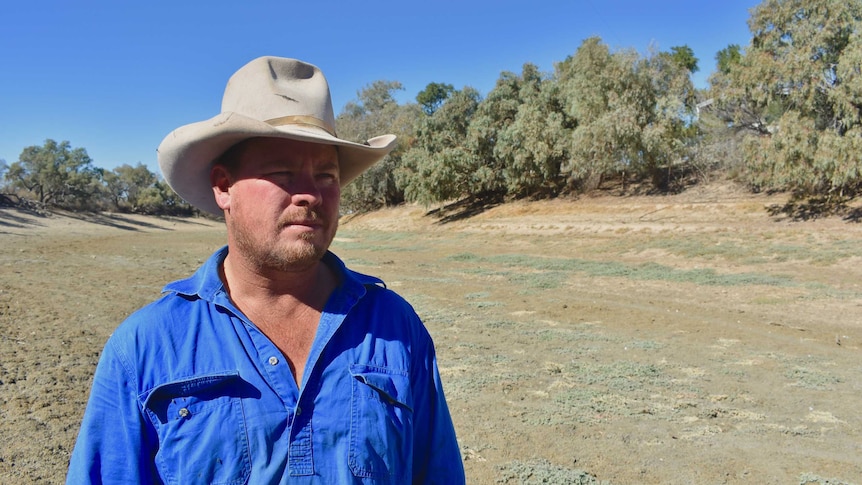 A man in a well-worn wide-brimmed hat and blue shirt stands in the middle of a dry creek bed with trees in the background