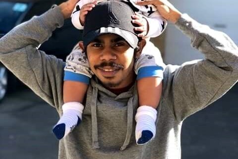Man wearing a greay jumper and black cap, holding a young smiling child on his shoulders