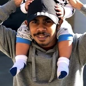 Man wearing a greay jumper and black cap, holding a young smiling child on his shoulders