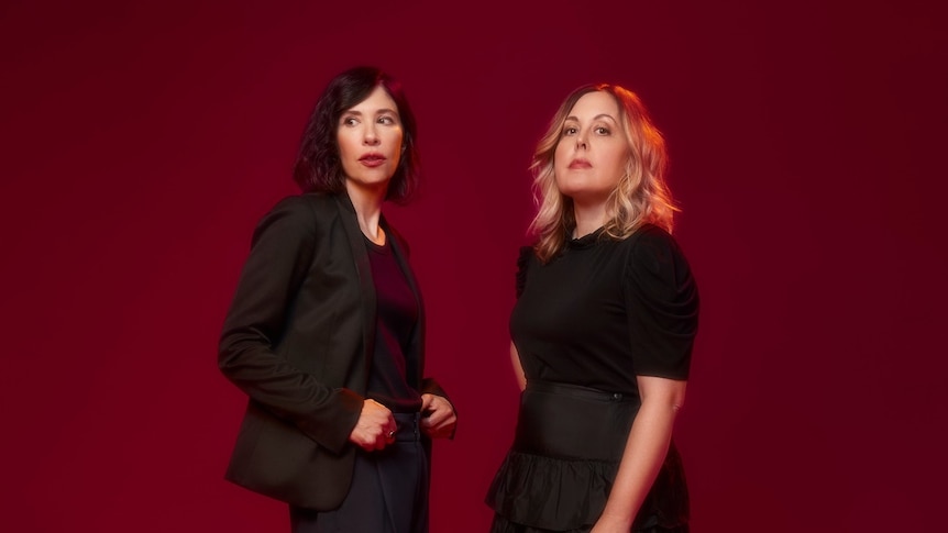 two women wearing black clothing stand before a maroon background