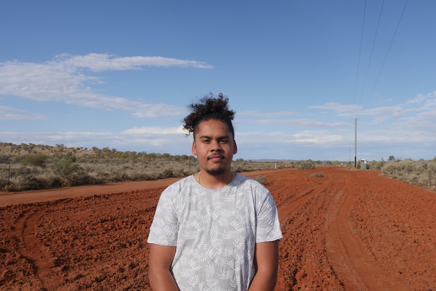 A young man stands on an outback road in a white shirt