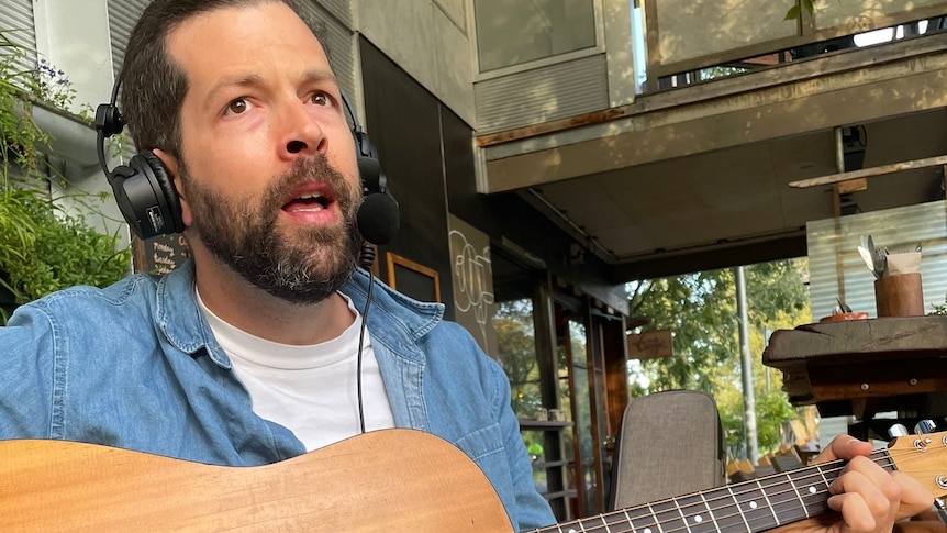 man with beard and headphones on holding a guitar singing outside a cafe