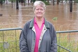 Sandy Willcott, pink shirt, jacket, standing in front of fence and grass, brown floodwater behind.