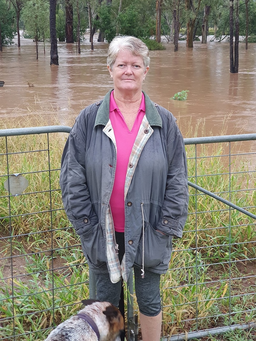 Sandy Willcott, pink shirt, jacket, standing in front of fence and grass, brown floodwater behind.