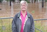 Sandy Willcott, pink shirt, jacket, standing in front of fence and grass, brown floodwater behind.